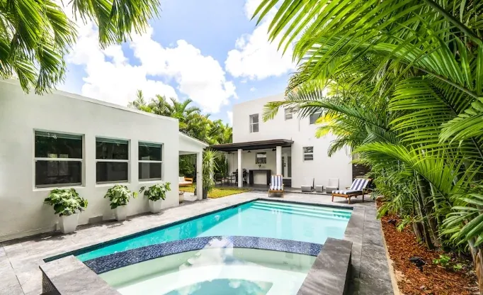 A backyard pool and spa beside a two-story white house framed by palms